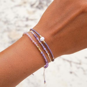 May include: Three delicate bracelets with purple beads and gold accents are displayed on a wrist. One bracelet features a pearl. The bracelets are strung on a thin, off-white cord. The background is a light, speckled surface.