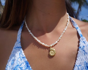 May include: A white pearl necklace with a gold ammonite pendant. The necklace is worn by a person with a blue and white floral patterned top.