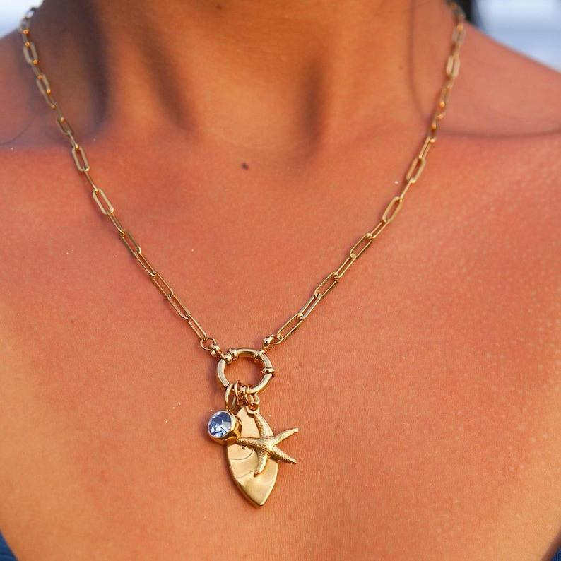 K&ouml;nnte beinhalten: Eine goldene Kettenkette mit einem Anh&auml;nger in Form eines Surfbretts, eines Seesterns und eines blauen Edelsteins. Die Kette ist aus einer Gliederkette gefertigt.