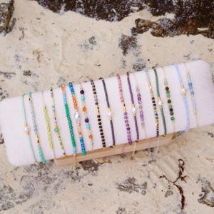 May include: A collection of delicate beaded bracelets in various colors, including green, blue, purple, and gold. Each bracelet features small beads and is displayed on a white rectangular stand. The bracelets are presented on a sandy surface.