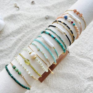 May include: A display of various beaded bracelets in shades of green, blue, pink, and gold. The bracelets feature gemstones, pearls, and gold chains. Some bracelets have a single large pearl or stone, while others are made of small beads.