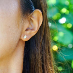 Puede incluir: Primer plano de una oreja con un pequeño pendiente de botón redondo de oro. El pendiente tiene un diseño simple y minimalista. El fondo está borroso, con bokeh verde y amarillo.