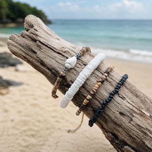 May include: A set of four bracelets featuring a variety of beads. One bracelet has a white shell and metallic accents, one has flat white disc beads, one has small brown wooden beads, and one is composed of small black beads with a knotted cord finish.