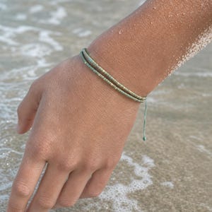 Op de afbeelding: Een dubbelstrengs armband met kleine, cilindervormige saliegroene kralen met metallic gouden accenten. De armband is gemaakt van een dun, bijpassend saliegroen koord en heeft een verstelbare schuifknoopsluiting met twee losse uiteinden.