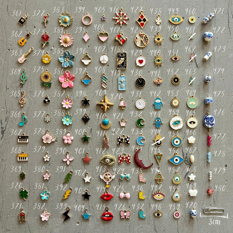 K&ouml;nnte beinhalten: Eine Sammlung von 100 kleinen Charms, jeder mit einem anderen Design. Die Charms sind aus Metall und haben eine Vielzahl von Farben, darunter Gold, Silber, Blau, Rot, Gr&uuml;n und Pink. Die Charms sind in einem Rastermuster auf einer grauen Oberfl&auml;che angeordnet. Die Charms sind von 361 bis 480 nummeriert.