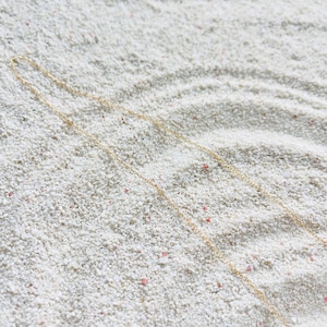 May include: A delicate gold necklace with a fine chain, resting on a bed of white sand. The necklace is simple and elegant, with a subtle shine. The sand has small pink and red flecks.
