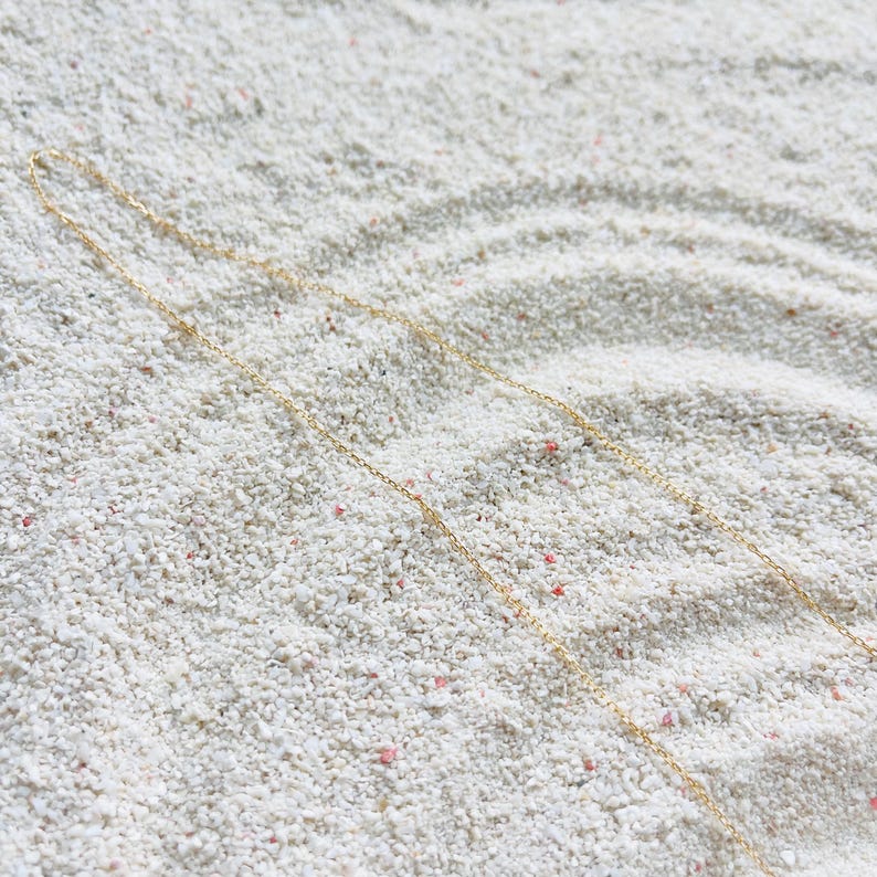 May include: A delicate gold necklace with a fine chain, resting on a bed of white sand. The necklace is simple and elegant, with a subtle shine. The sand has small pink and red flecks.