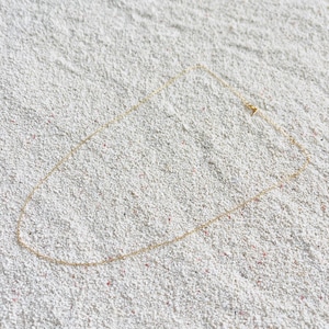 May include: A delicate gold-colored necklace with a simple chain design. The necklace is laid on a textured, white surface. The chain is thin and appears to be approximately 18 inches long.