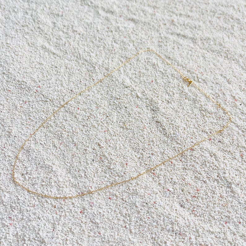 May include: A delicate gold-colored necklace with a simple chain design. The necklace is laid on a textured, white surface. The chain is thin and appears to be approximately 18 inches long.