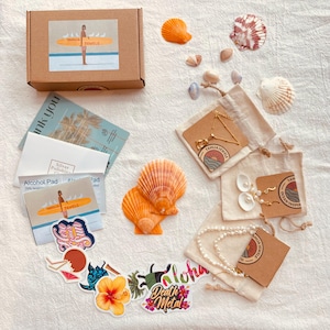 May include: An overhead shot of jewelry and accessories, including a brown box with a logo, stickers, seashells, and jewelry pouches. The jewelry includes a gold necklace and pearl necklace. The stickers have the words "Aloha" and "Death Metal".