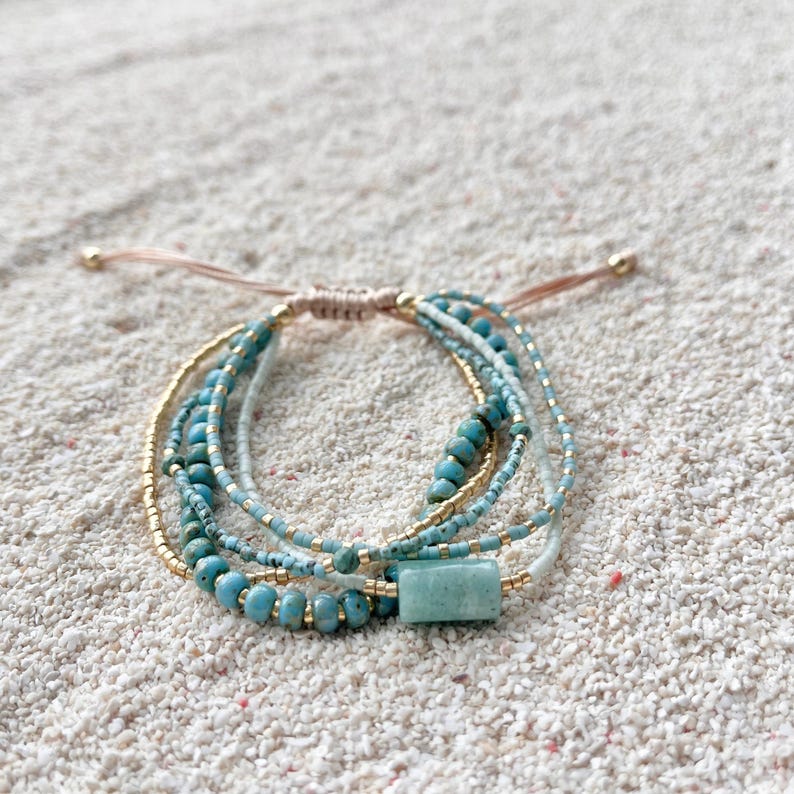 May include: A multi-strand bracelet featuring turquoise-colored beads, small metallic gold-tone beads, and a single rectangular light green stone bead. The bracelet includes adjustable tan cord ties with small gold-tone beads at the ends.