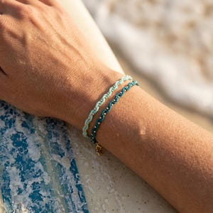 May include: Two braided bracelets in shades of teal and seafoam green are worn on a wrist. The bracelets have a rope-like design with small, gold-colored beads. A small gold charm hangs from the bottom bracelet. The background is blurred, suggesting a beach setting.