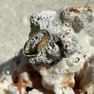Op de afbeelding: Een goudkleurige ring met een zonnesterontwerp. De ring ligt op een stuk koraal. Het zonnesterontwerp bevindt zich aan de bovenkant van de ring, met een gebogen band die de vinger omsluit.