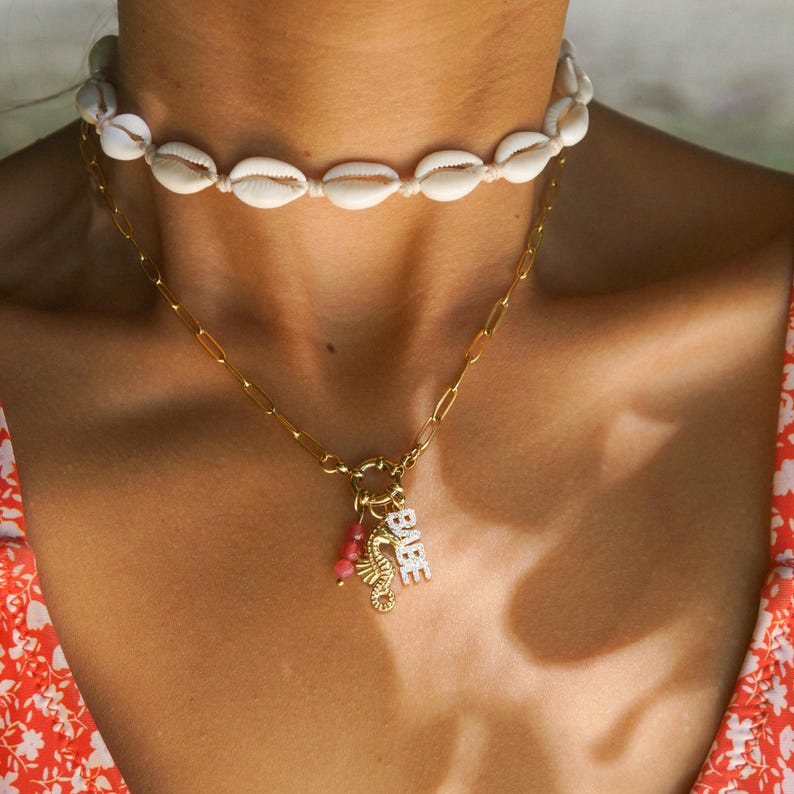 K&ouml;nnte beinhalten: Eine goldene Kettenkette mit einem Anh&auml;nger, auf dem "BAE" steht, und einem Seepferdchen-Anh&auml;nger. Die Kette wird &uuml;ber einem wei&szlig;en Muschel-Choker getragen.