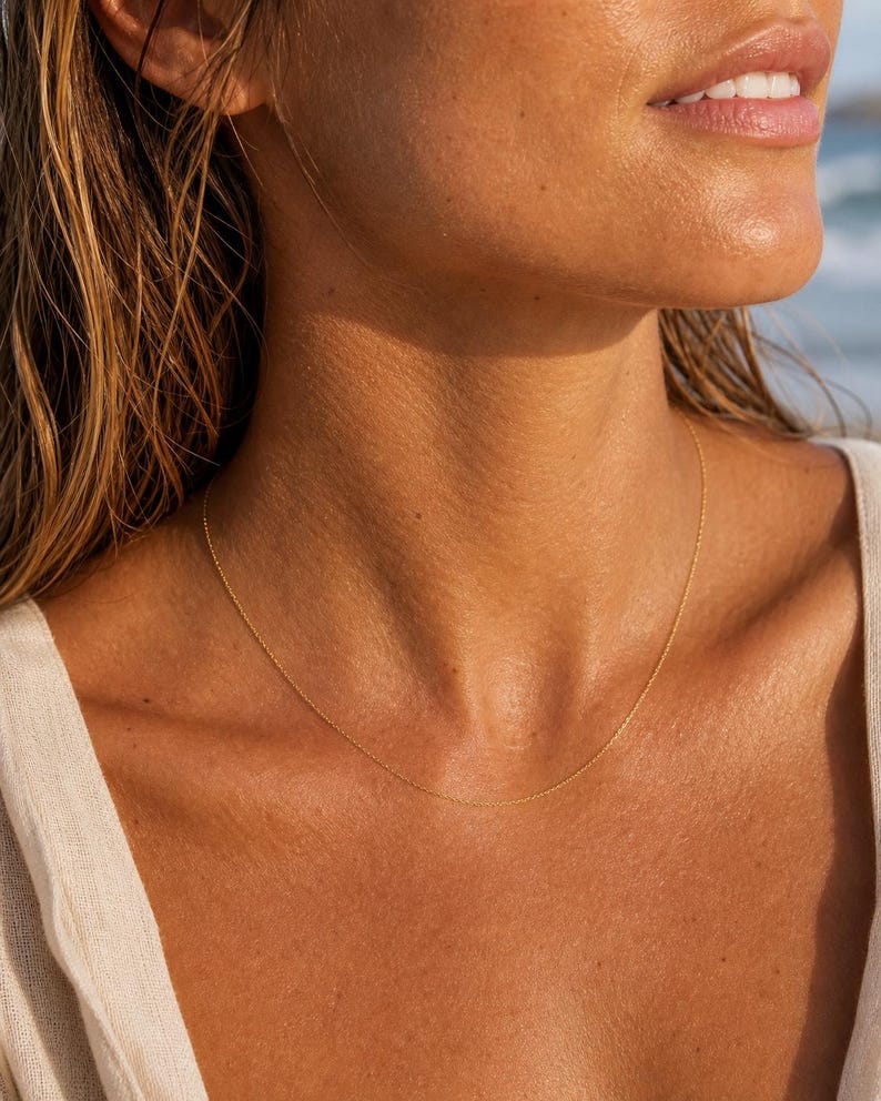 May include: A delicate gold necklace resting on a person's skin. The fine chain is a simple, elegant design. The background is blurred, suggesting a beach setting, with sunlight reflecting off the skin.