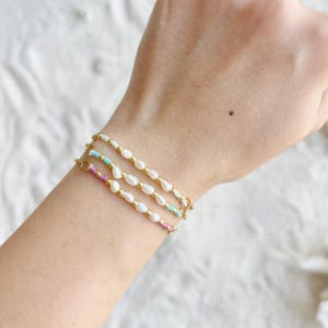May include: Three gold bracelets, each featuring white pearl beads and colourful turquoise and pink accents. The bracelets are shown on a wrist, highlighting their elegant design. These bracelets are ideal for adding a touch of sophistication to any outfit.