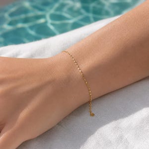Könnte beinhalten: Ein zartes Goldkettenarmband mit kleinen, flachen, reflektierenden runden Gliedern, die das Licht einfangen. Das Armband hat ein dünnes Design mit einem kleinen runden Anhänger am Ende der verstellbaren Verlängerungskette für einen dezenten Glanz.