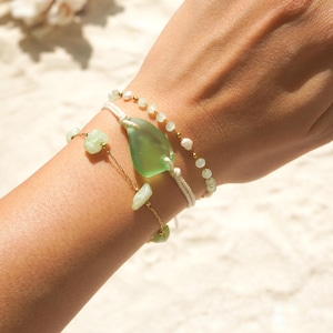 May include: Three bracelets are shown on a wrist. One has a large, green, leaf-shaped glass bead. Another has small, white beads. The third has small, green, irregular-shaped beads strung on a gold chain.