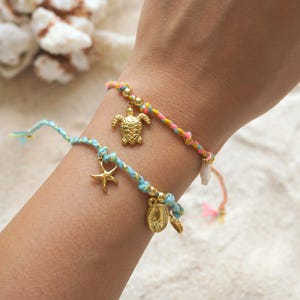 May include: Two colourful woven bracelets with gold charms are displayed on a wrist. One bracelet is blue and the other is multi-coloured. The charms include a turtle, a starfish, and a coin. The bracelets are a beach-themed accessory.