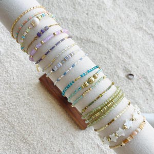 May include: A selection of delicate beaded bracelets in various colours, including gold, blue, green, and purple. Some bracelets feature small beads, while others have larger stones or star-shaped accents. The bracelets are displayed on a white cylindrical stand.