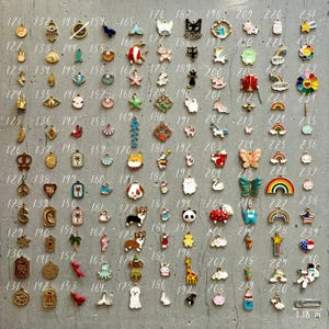 K&ouml;nnte beinhalten: Eine Sammlung von 100 Emaille-Anh&auml;ngern in verschiedenen Farben und Designs. Die Anh&auml;nger sind in Reihen auf einer grauen Oberfl&auml;che angeordnet. Die Anh&auml;nger umfassen Tiere, Pflanzen, Lebensmittel und andere Gegenst&auml;nde. Die Anh&auml;nger sind etwa 3 cm gro&szlig;.