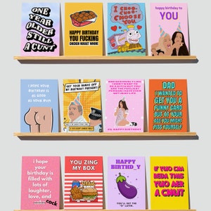 May include: A collection of humorous birthday cards displayed on a wooden shelf. The cards feature various designs, including cartoon illustrations, bold text, and playful messages. The color palette is vibrant, with shades of blue, pink, yellow, and orange.