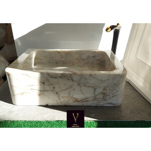 May include: A white marble sink with brown veining. The sink is rectangular and has a slightly rounded edge. It is sitting on a gray surface.