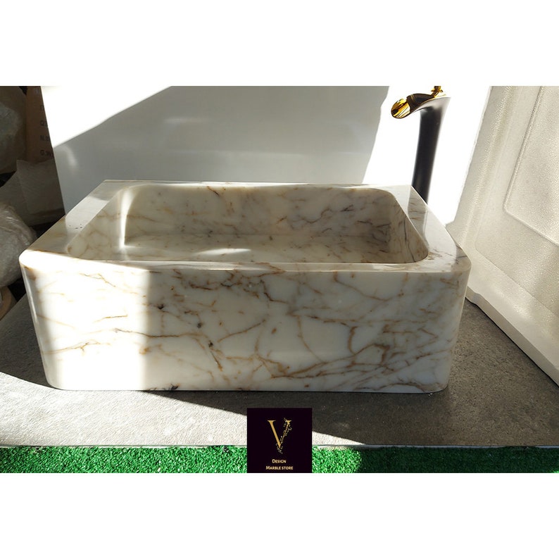 May include: A white marble sink with brown veining. The sink is rectangular and has a slightly rounded edge. It is sitting on a gray surface.