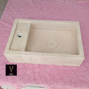 May include: Beige stone rectangular sink with a built-in drainboard and a circular drain. The sink features a rectangular basin and a small, round drain hole on the drainboard. The sink is made of light-coloured stone.