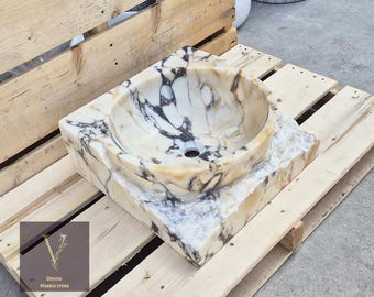 May include: A square, white marble sink with brown and gray veining. The sink is set on a wooden pallet.