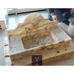 Op de afbeelding: Een rechthoekige marmeren wastafel met een gebogen backsplash. De wastafel heeft een lichtbeige en bruin geaderd patroon. De wastafel staat op een houten pallet. Het logo "V Design Marble Store" staat in de onderste hoek.