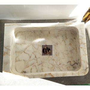 May include: A white marble sink with gold veining. The sink is rectangular and has a rounded edge. The sink is set into a white countertop.