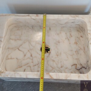 May include: A white marble bathroom sink with a gold drain stopper. The sink is 16 inches wide.