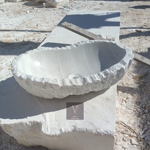May include: A white marble sink with a rough, textured exterior. The sink is oval-shaped and has a smooth, polished interior. The sink is sitting on a block of white marble.