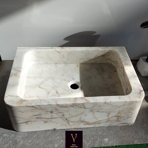 May include: A white marble bathroom sink with a rectangular shape and a rounded edge. The sink has a drain hole in the center and a subtle veining pattern.