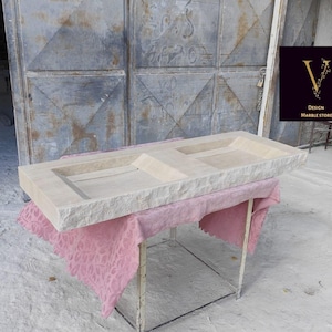 Peut inclure: Un évier rectangulaire en pierre beige clair avec deux bassins encastrés. L'évier est présenté sur un support métallique recouvert d'un tissu rose à motifs. L'arrière-plan présente une porte en métal vieilli et un logo "Design Marble Store".