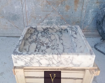 May include: A white marble sink with gray veining, sitting on a wooden crate. The sink is square shaped with a rectangular cutout for the basin. The crate has the text "Design Marble Store" on it.