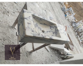 May include: A white marble sink with gray veining, mounted on a wooden stand. The sink is rectangular with a rounded edge and a single drain hole.