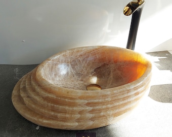 Könnte beinhalten: Ein beigefarbenes Onyx-Waschbecken mit einem geschichteten Design. Das Waschbecken hat einen goldfarbenen Abfluss und wird mit einem schwarzen und goldenen Wasserhahn kombiniert.