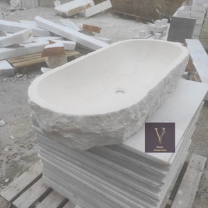 Könnte beinhalten: Eine Badewanne aus weißem Stein mit einer rauen, strukturierten Außenseite und einem glatten Innenraum. Die ovale Wanne steht auf einem Stapel weißer Steinplatten. Das Bild enthält den Text "DESIGN MARBLE STORE".