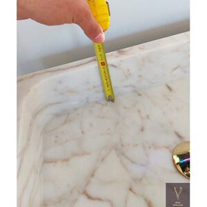 May include: A white marble sink with a gold drain cover. A measuring tape is used to measure the sink's dimensions, showing a measurement of 15 inches.