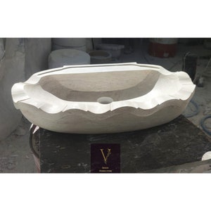 May include: A light-coloured, carved stone sink with a scalloped edge design. The oval-shaped basin features a central drain hole and a smooth interior. The sink is displayed on a dark surface, showcasing the natural stone texture. The logo "V Design Marble Store" is visible.