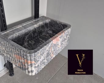May include: A rectangular marble sink with a fluted exterior. The sink is primarily gray with black and pink veining. The sink is mounted on a black metal support. The logo for Design Marble Store is in the lower right corner.
