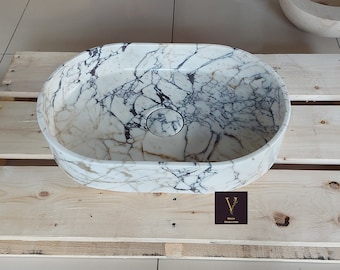 May include: Oval-shaped white marble sink with a gray and brown veining pattern. The sink has a round drain hole in the center.