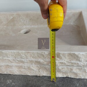 May include: A close-up of a rectangular stone sink with a rounded edge. A measuring tape is used to measure the width of the sink, which is 15 inches.