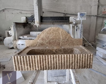 May include: A large, brown marble bathroom sink with a fluted front and a curved backsplash. The sink is in a workshop setting, with a CNC machine in the background. The sink is supported by a wooden frame.