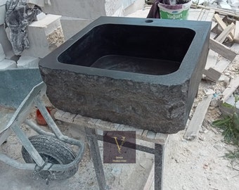 May include: A black granite sink with a rough, textured exterior. The sink is rectangular and has a single drain hole. It is sitting on a wooden stand.