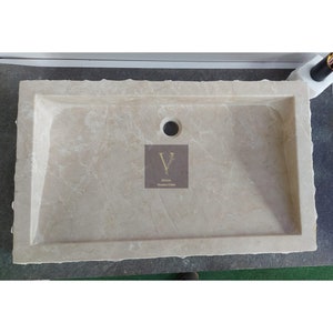 May include: A rectangular, off-white marble sink with a single drain hole. The sink has a rough, uneven edge and a smooth, polished interior.