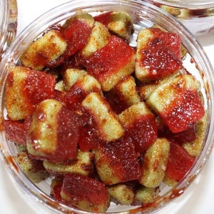 Puede incluir: Un tarro de vidrio transparente lleno de caramelos gomosos rojos y verdes. Los caramelos están cubiertos con una salsa roja y tienen una textura ligeramente granulada.