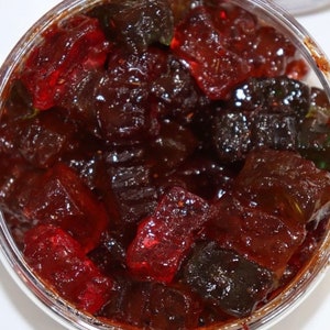 Puede incluir: Un tarro de vidrio transparente lleno de fruta confitada roja, verde y negra. La fruta está cortada en cuadrados pequeños y está cubierta con un glaseado brillante.